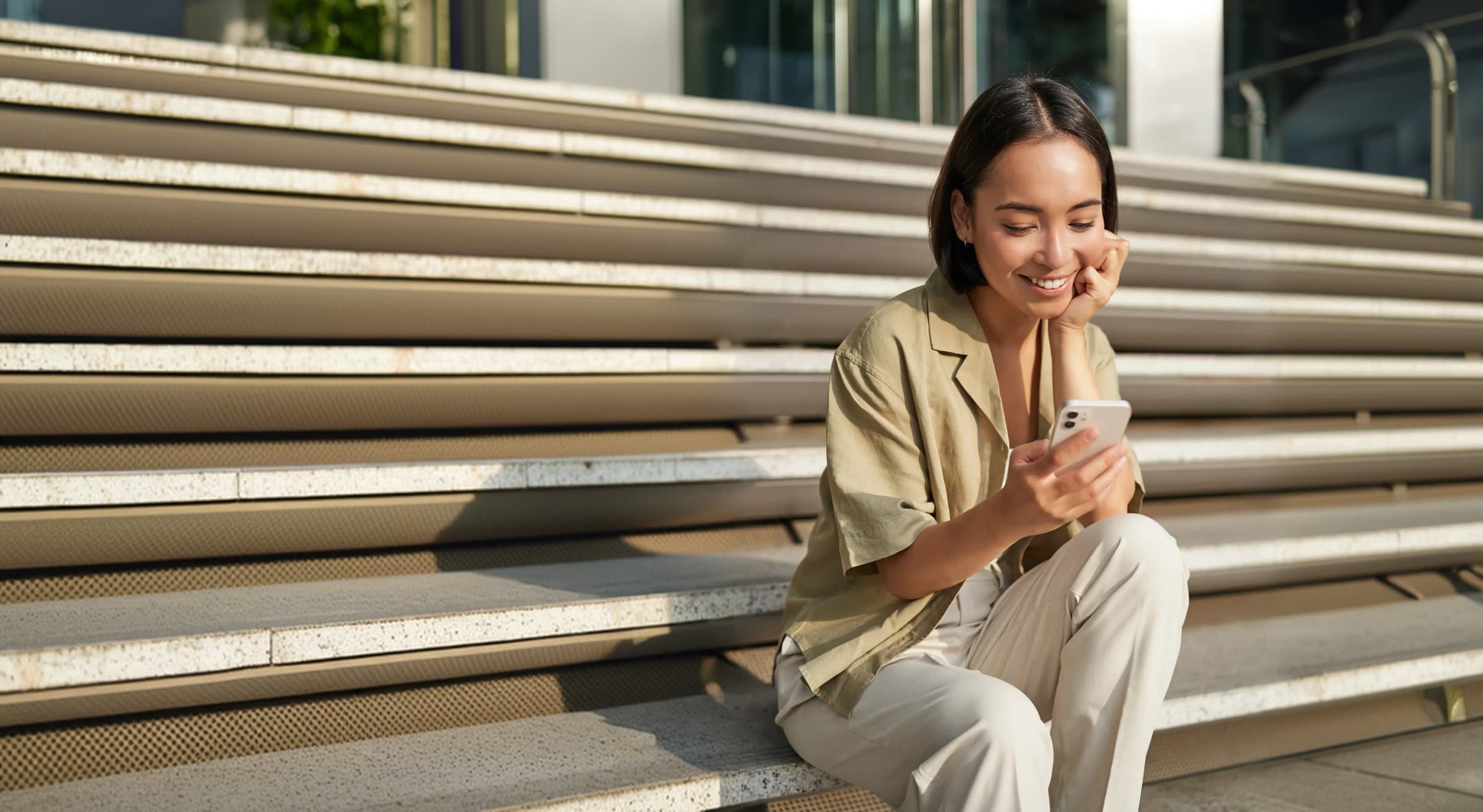 A smiling woman sitting on city steps, checking her investments on her phone